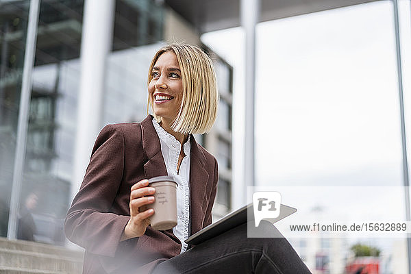 Lächelnde junge Geschäftsfrau mit Tablette und Kaffee zum Mitnehmen in der Stadt