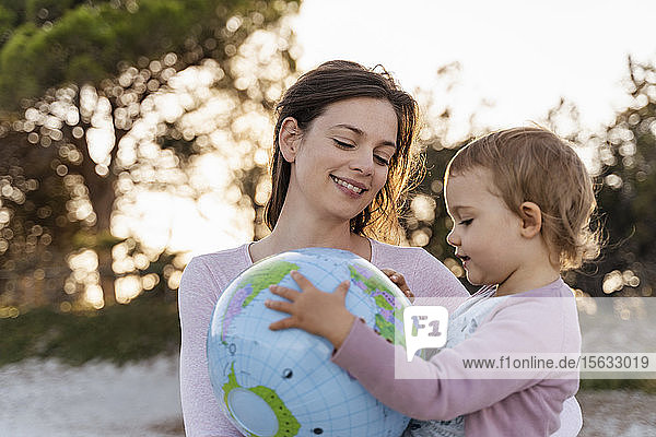 Porträt einer glücklichen Mutter  die zusammen mit ihrer kleinen Tochter auf den Strandball der Erde schaut