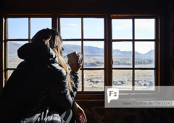 Frau trinkt eine Tasse Kaffee  während sie durch das Fenster die Berge bewundert