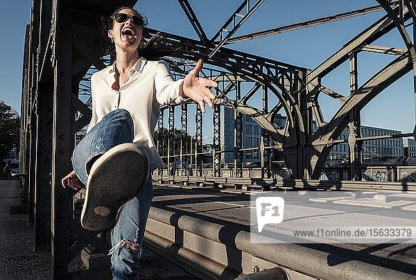 Young woman with sunglasses kicking in the air on a bridge in the evening light