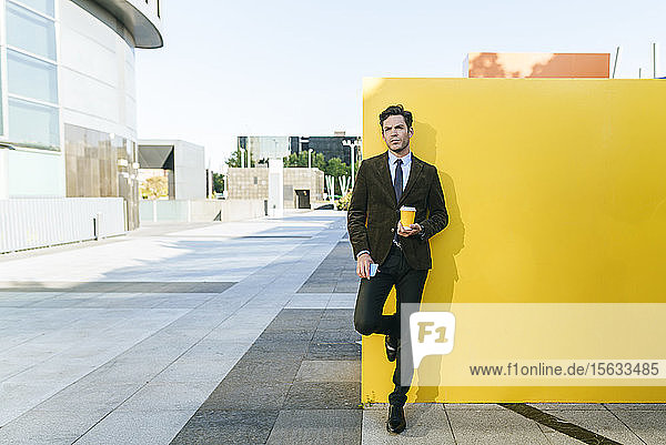 Geschäftsmann mit Kaffee zum Mitnehmen und Telefon neben der gelben Wand im städtischen Geschäftsviertel  Madrid  Spanien