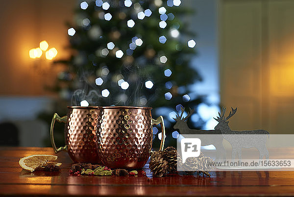 Nahaufnahme von Glühwein mit Essen und Dekorationen auf dem Tisch vor beleuchtetem Weihnachtsbaum zu Hause