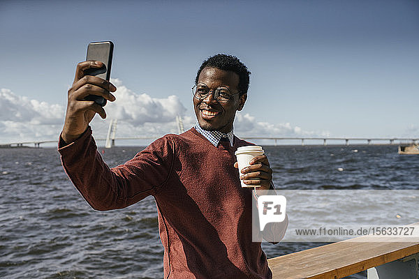 Junger Mann mit Smartphone-Selfie am Meer  hält Kaffee zum Mitnehmen in der Hand