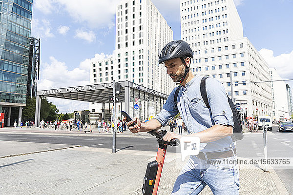 Geschäftsmann betrachtet das Smartphone beim Pendeln auf dem Elektroroller in der Stadt  Berlin  Deutschland