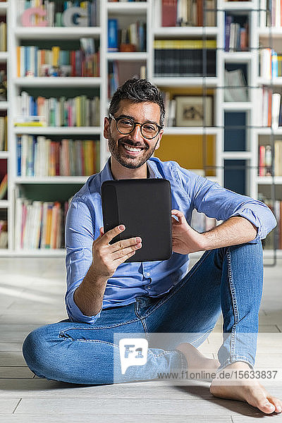 Porträt eines barfüssigen jungen Mannes  der vor Bücherregalen auf dem Boden sitzt und ein digitales Tablett benutzt