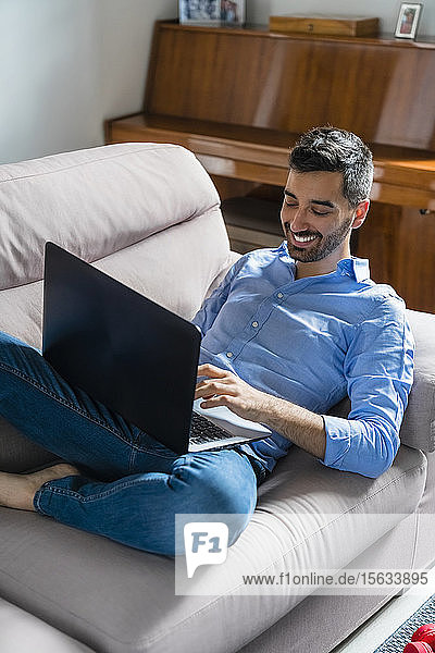 Smiling young man sitting on the couch at home using laptop