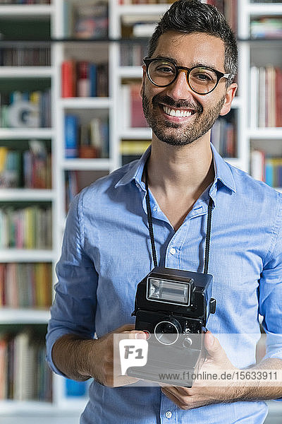 Porträt eines glücklichen jungen Mannes mit Sofortbildkamera vor einem Bücherregal stehend