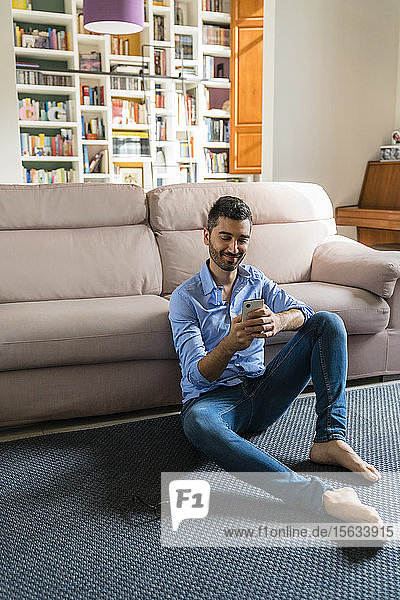 Porträt eines lächelnden jungen Mannes  der zu Hause auf dem Boden des Wohnzimmers sitzt und ein Smartphone benutzt