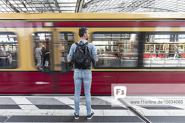 Rückansicht eines Mannes mit Rucksack auf dem Bahnsteig während der Einfahrt des Zuges