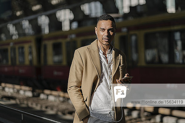 Portrait of man standing at train station using smartphone and earphones  Berlin  Germany