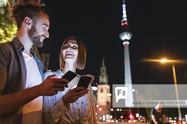 Glückliches Paar benutzt nachts in der Stadt Smartphones  Fernsehturm im Hintergrund  Berlin  Deutschland