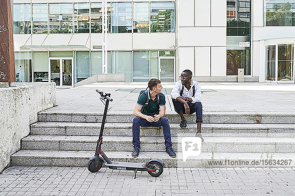 Zwei zufällige Geschäftsleute unterhalten sich auf einer Treppe in der Stadt neben einem E-Scooter