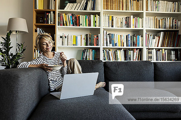 Reife Frau,  die einen Laptop benutzt und zu Hause auf der Couch zu Mittag isst