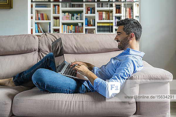 Lächelnder junger Mann  der zu Hause auf der Couch liegt und seinen Laptop benutzt
