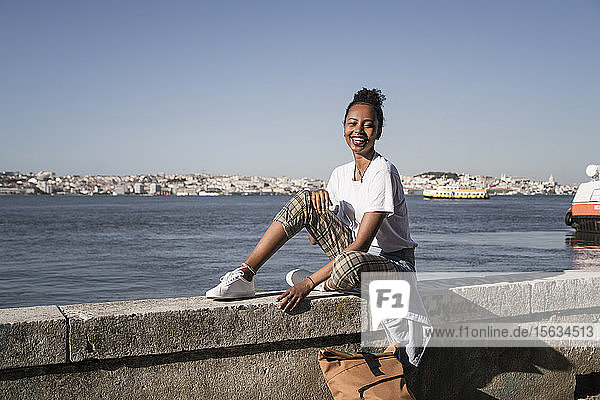 Portrait of happy young woman sitting on a wall at the waterfront  Lisbon  Portugal