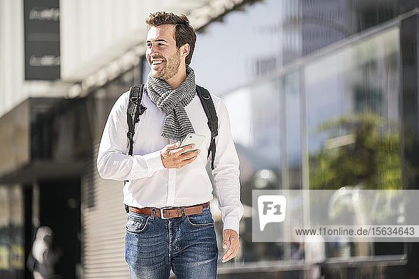 Lächelnder junger Mann mit Rucksack und Handy in der Stadt unterwegs