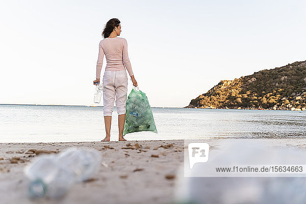 Rückenansicht einer Frau  die mit einem Müllsack mit gesammelten leeren Plastikflaschen am Strand steht