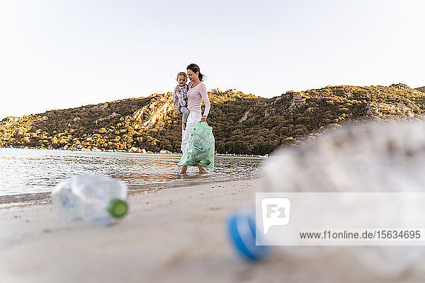 Frau trägt kleine Tochter auf dem Arm und sammelt leere Plastikflaschen am Meeresufer