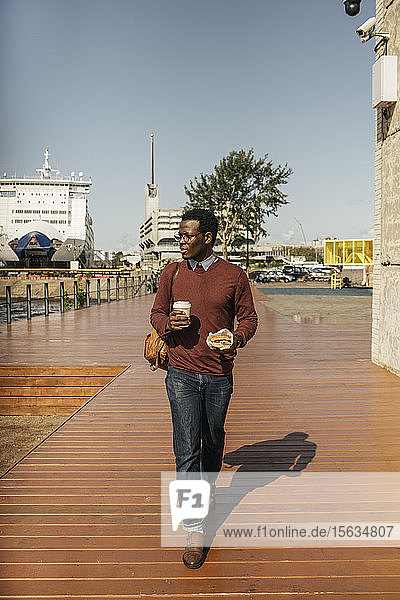 Junger Mann mit Kaffeetasse und Hamburger beim Spaziergang am Hafen
