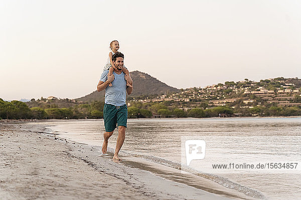 Glücklicher Vater trägt seine Tochter bei Sonnenuntergang am Strand auf den Schultern