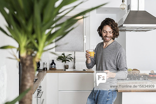 Mann hält zu Hause in der Küche ein Glas Orangensaft und ein Handy