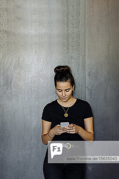 Porträt einer ungezwungenen jungen Geschäftsfrau  die ein Smartphone vor einer grauen Wand benutzt