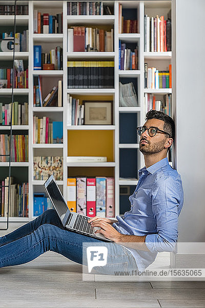 Porträt eines nachdenklichen jungen Mannes  der mit seinem Laptop vor Bücherregalen auf dem Boden sitzt