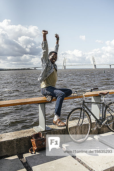 Junger Mann mit Fahrrad  sitzt auf einer Reling am Meer und streckt sich