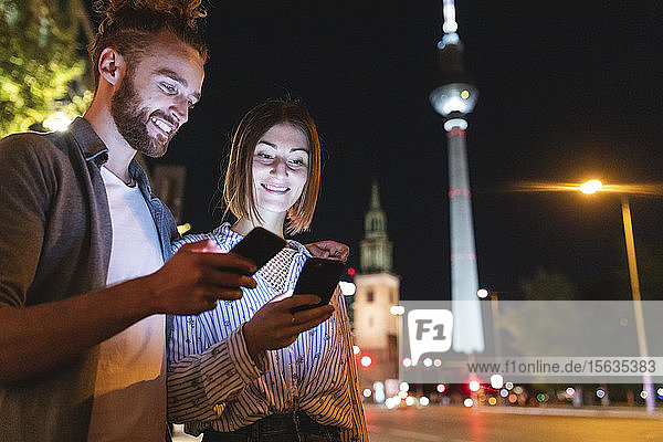 happy couple using smartphones in the city at night  Fernsehturm in the background  Berlin  Germany