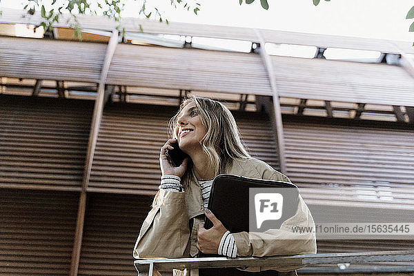 Junge blonde Frau benutzt ein Smartphone und hält eine Laptop-Tasche