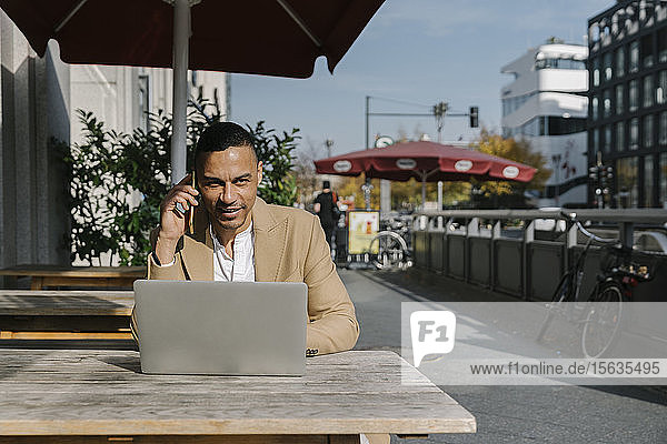 Porträt eines lächelnden Geschäftsmannes am Telefon  der mit seinem Laptop in einem Straßencafé sitzt