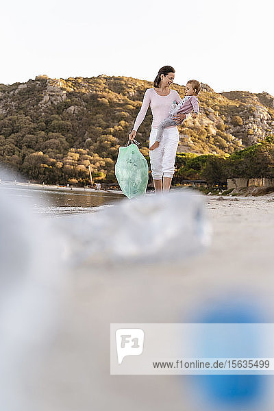 Frau mit kleiner Tochter am Arm sammelt leere Plastikflaschen am Strand