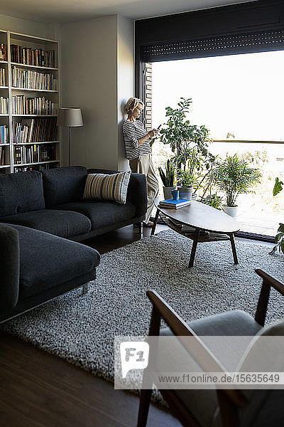 Reife Frau  die zu Hause am Fenster im Wohnzimmer steht und ein Smartphone benutzt