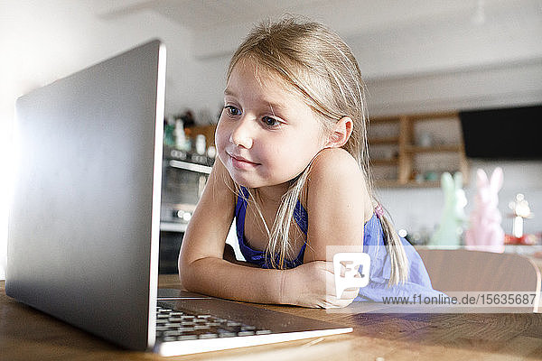 Portrait of little girl leaning on kitchen table at home looking at laptop