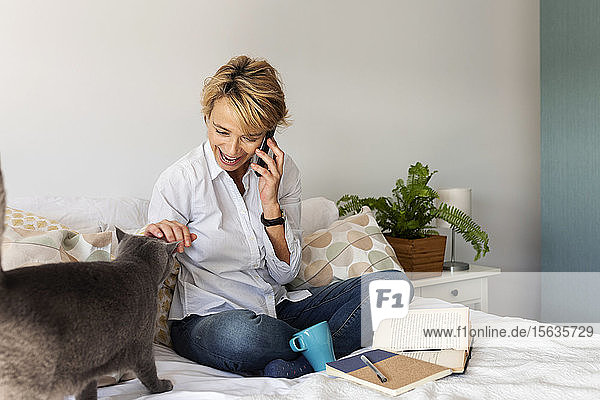 Reife Frau mit Katze sitzt zu Hause auf dem Bett und telefoniert