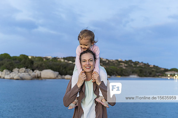 Porträt einer Mutter  die ihre Tochter bei Sonnenuntergang an der Strandpromenade auf den Schultern trägt