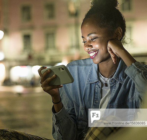 Porträt einer glücklichen jungen Frau  die nachts auf ein Smartphone schaut  Lissabon  Portugal