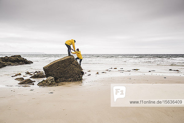 Junge Frau in gelber Regenjacke beim Klettern auf einem Felsen am Strand  Bretagne  Frankreich