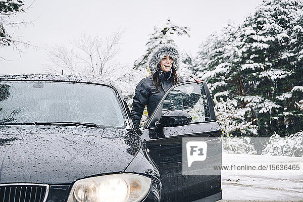 Lächelnde junge Frau steht neben geparktem Auto im Winterwald und schaut in die Ferne
