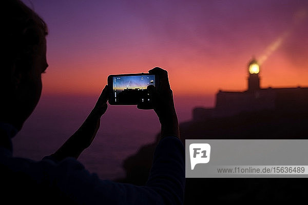 Portugal  Algarve  Über-Schulter-Ansicht einer Person  die in der Morgendämmerung per Smartphone Fotos vom Leuchtturm von Cape Saint Vincent macht