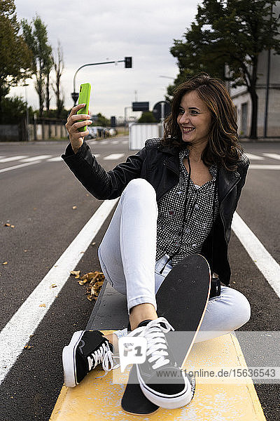 Lächelnde junge Frau mit Skateboard auf einem Poller sitzend  die mit einem Smartphone Selbsthilfe nimmt