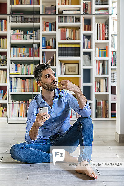 Porträt eines nachdenklichen jungen Mannes mit Smartphone  der zu Hause vor Bücherregalen auf dem Boden sitzt und auf Distanz schaut