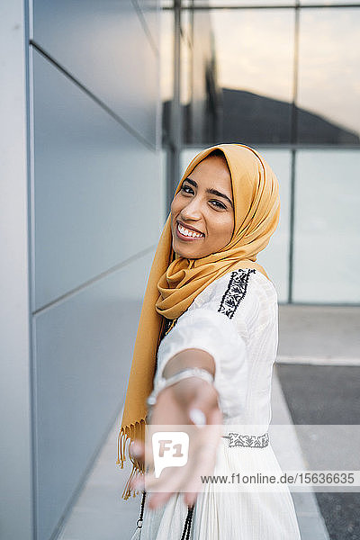 Junge muslimische Frau lächelt  trägt gelben Hidschab und reicht ihre Hand