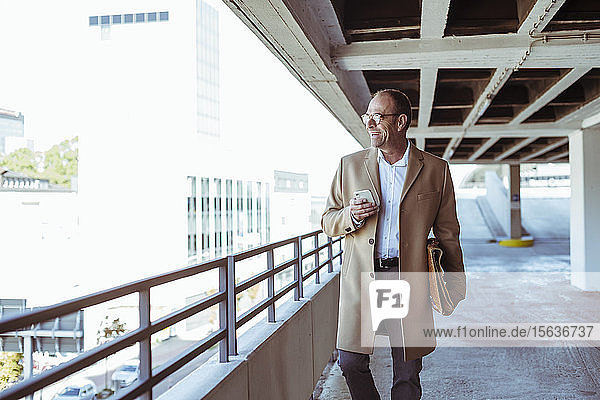 Happy mature businessman with smartphone and briefcase walking on parking deck