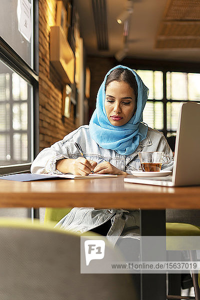 Businesswoman wearing turquoise hijab in a cafe and writing in notebook
