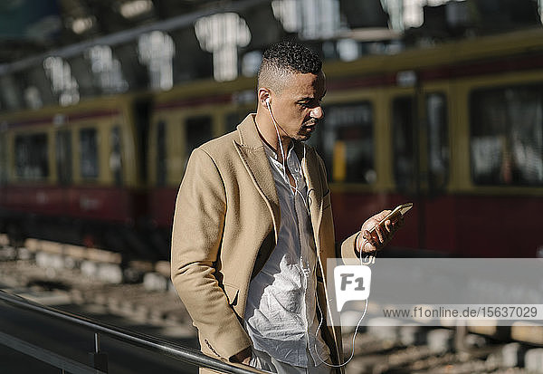 Man standing at train station using smartphone and earphones  Berlin  Germany