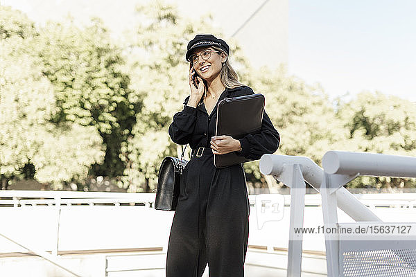 Junge blonde Geschäftsfrau mit schwarzem Hut  die eine Laptop-Tasche hält und ein Smartphone benutzt
