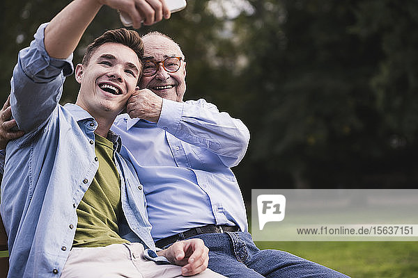 Älterer Mann und Enkel sitzen zusammen auf einer Parkbank und nehmen Selfie mit Smartphone