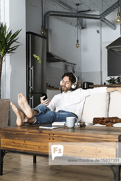 Entspannter Mann sitzt zu Hause auf der Couch mit Handy und Kopfhörern