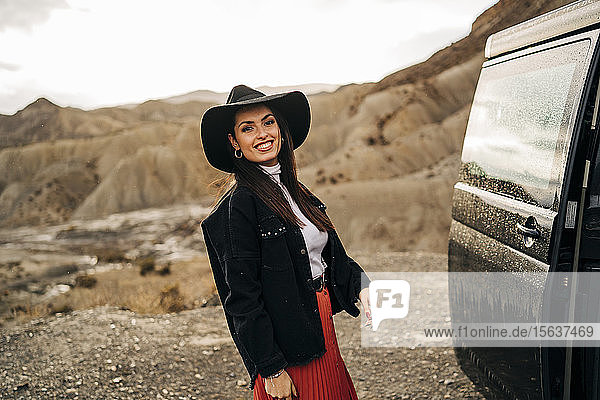 Portait of smiling young woman in desert landscape standing next to camper van  Almeria  Andalusia  Spain